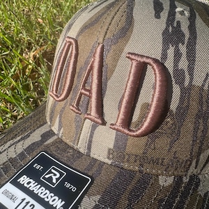 May include: Camouflage trucker hat with the word "DAD" embroidered in brown. The hat features a curved brim and an adjustable strap. The Richardson 112 Trucker hat is visible on the label.