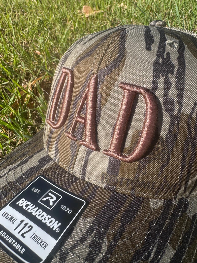May include: Camouflage trucker hat with the word "DAD" embroidered in brown. The hat features a curved brim and an adjustable strap. The Richardson 112 Trucker hat is visible on the label.
