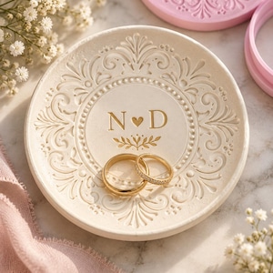 May include: A round, off-white ceramic ring dish featuring an embossed scrollwork border and the gold-printed letters N, a heart symbol, and D in the center. Two gold-toned rings, one plain and one with small stones, rest inside the dish.