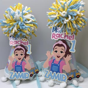 Puede incluir: Dos sombreros de fiesta de cumpleaños con una imagen de dibujos animados de una niña, el número 1 y el nombre "Ms. Rachel". Cada sombrero tiene una cinta azul y un pompón en amarillo, blanco y azul. El nombre "ZAMID" también está impreso en los sombreros.