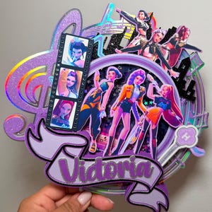 Pode incluir: Um topo de bolo multicamadas com um grupo de figuras estilizadas em trajes vibrantes. O design inclui uma tira de filme com retratos, uma nota musical e o nome "Vitoria" em uma faixa. O esquema de cores é predominantemente roxo, com detalhes holográficos e brilhantes.