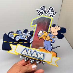 May include: A colorful birthday cake topper featuring a race car, Mickey Mouse in a racing suit, the number 1, and the name "ALAN". The topper includes checkered flags and a road design.