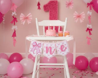 May include: A pink-themed first birthday party setup. A white high chair is decorated with a banner that reads "one". A small cake with candles and cupcakes sit on the chair. Pink balloons and decorations complete the festive scene.