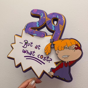 Könnte beinhalten: Dekoratives Schild mit der Zahl 29 in Lila mit einem bunten Muster. Das Schild enthält eine Zeichentrickfigur und eine Sprechblase mit dem Text "...But at what cost?" in Lila.