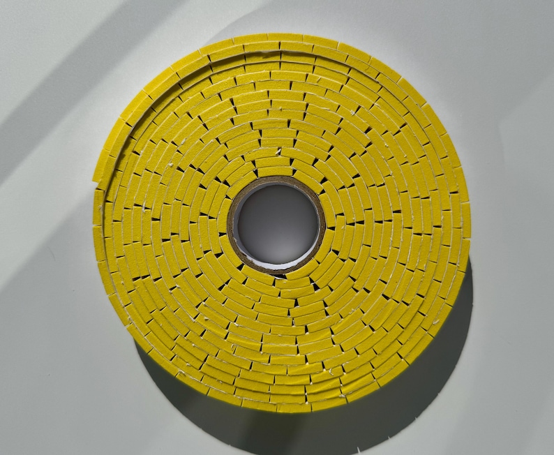 May include: A yellow roll of tape with a circular shape. The tape is made up of small, rectangular pieces that are stacked on top of each other.