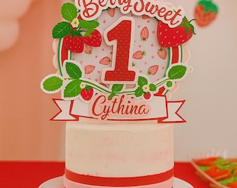 May include: A white frosted cake with red and pink stripes, topped with a "Berry Sweet" cake topper. The topper features strawberries, green leaves, and the number "1" with the name "Cythina". The cake sits on a white pedestal.