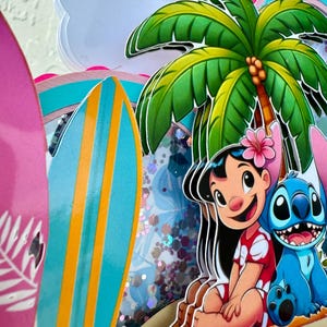 May include: A layered papercraft with a tropical theme. It features a palm tree, surfboards, and cartoon characters Lilo and Stitch. The colour scheme is vibrant, with shades of pink, blue, green, and yellow.