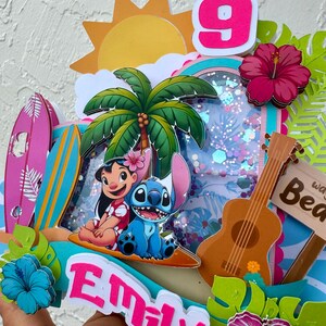 May include: A vibrant, layered birthday decoration with Lilo and Stitch, a palm tree, a ukulele, and a sign that says "Beach". The number 9 is displayed, along with the name "Emily". Decorative hibiscus flowers and surfboards complete the tropical theme.