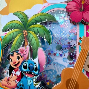 May include: A vibrant, layered papercraft featuring cartoon characters Lilo and Stitch, a palm tree, a ukulele, and a hibiscus flower. The design includes a sunny sky, a rainbow, and a shaker element with glitter. The overall theme is tropical and celebratory.