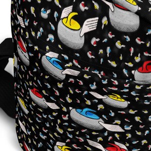May include: Black crossbody bag featuring a repeating pattern of curling stones in red, yellow, and blue. The bag has a black zip and strap. The curling stones have white wings against a background of colourful flecks.