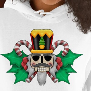 May include: A selection of hooded sweatshirts in white, black, pink, light blue, and grey. Each sweatshirt displays a graphic of a nutcracker with a moustache, a top hat, and candy cane accents, surrounded by green holly leaves.