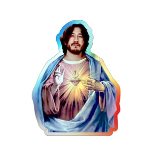 Könnte beinhalten: Ein Aufkleber mit einem Mann im Stil Jesu, mit Heiligenschein und leuchtendem Herzen. Er trägt eine hellrosa Tunika und ein blaues Gewand. Der Aufkleber hat einen holografischen, regenbogenfarbenen Rand.
