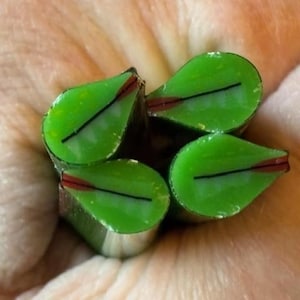 May include: Close-up of four green leaf-shaped glass beads. Each bead has a black line and red accent. The beads are held in a hand, with the background showing the skin's texture.