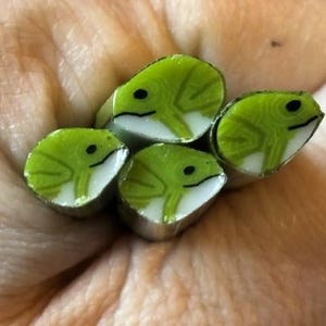 May include: Close-up of four green and white frog-shaped beads. Each bead features a detailed frog design with black eyes and a white belly. The beads are arranged together, showcasing their unique, handcrafted appearance.