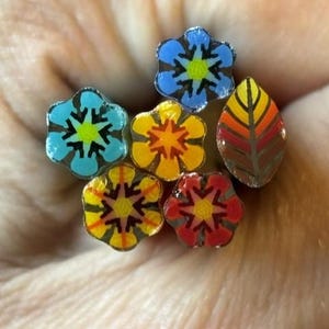 May include: A collection of six small polymer clay canes featuring intricate floral and leaf patterns. The designs include blue, yellow, orange, and red petals with geometric centres, alongside a single teardrop-shaped leaf cane with gradient stripes.