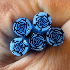 May include: Close-up of five blue cylindrical beads with a black spiral rose design on the top. The beads are held in a hand, and the background is a warm, neutral tone.