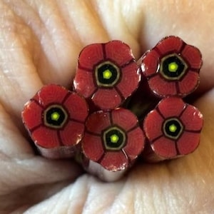 May include: Five cylindrical glass canes featuring a millefiori pattern of red flower petals surrounding a black hexagonal center with a bright yellow dot. The glass rods are arranged in a cluster, displaying a repeating floral motif in red, black, and yellow.