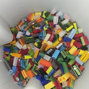 May include: A collection of colorful glass beads in various shapes and sizes, including cylindrical and rectangular forms. The beads are a mix of vibrant colors like red, yellow, green, blue, and orange. The beads are contained in a white container.