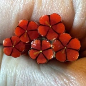May include: A cluster of five small, bright orange flower-shaped charms. Each charm features five rounded petals with dark brown lines radiating from a central yellow dot, creating a distinct floral pattern on each individual piece.