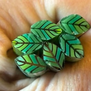 May include: A cluster of seven leaf-shaped beads. Each bead is a vibrant green with darker green and black vein details. The beads are held in a hand, showcasing their intricate design and color variations.