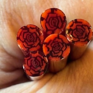 May include: Close-up of several orange and black polymer clay canes. Each cane end features a detailed rose design in black against an orange background. The canes are cylindrical and appear to be held in a hand.