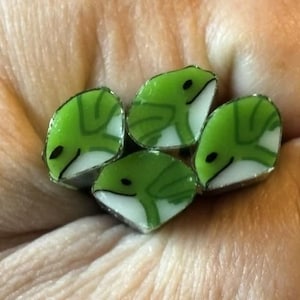 May include: A collection of four small, flat, triangular-shaped cabochons featuring a stylised green frog design on a white background. Each piece displays a minimalist graphic of a frog with a distinct green body and a white underbelly.