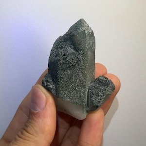 Puede incluir: Un grupo de cristales de color gris oscuro con una superficie rugosa y texturizada. El cristal tiene una parte superior puntiaguda y una base más clara y translúcida. El objeto se sostiene en una mano sobre un fondo blanco.