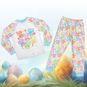 May include: Easter-themed pajama set featuring a long-sleeved white top with colorful bunny graphics and text, paired with matching pants. The sleeves and pants have a repeating pattern of pastel bunnies. The text includes words like "Risen" and "Faith".