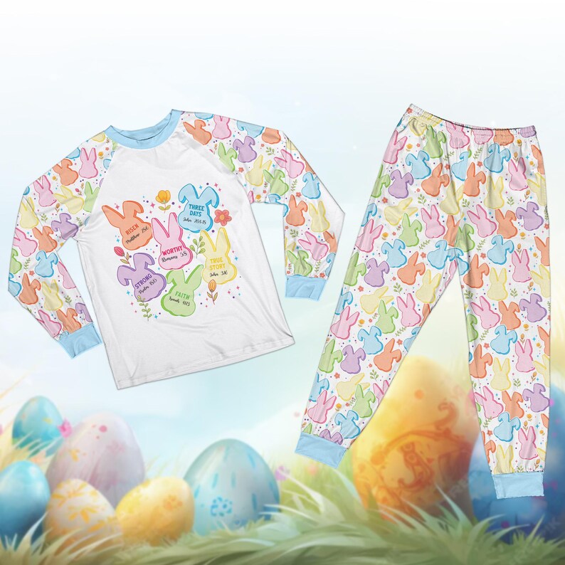 May include: Easter-themed pajama set featuring a long-sleeved white top with colorful bunny graphics and text, paired with matching pants. The sleeves and pants have a repeating pattern of pastel bunnies. The text includes words like "Risen" and "Faith".