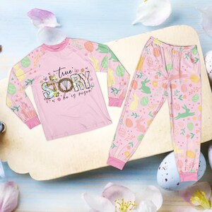 May include: Pink Easter-themed pajama set. The long-sleeved top features the text "True Story, he is risen" with floral and cross accents. The sleeves and pants are decorated with Easter eggs and bunnies.