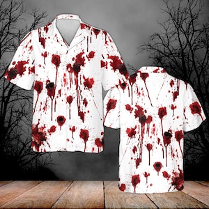 Puede incluir: Una camisa blanca de manga corta con botones, que presenta un estampado gráfico de salpicaduras de sangre roja y patrones de agujeros de bala oscuros en la parte delantera y trasera. La camisa tiene un cuello clásico y un dobladillo recto.