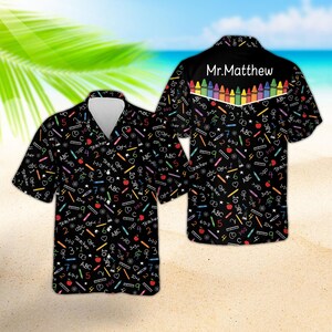 May include: A black short-sleeve button-down shirt featuring a colorful pattern of school supplies, numbers, and letters. The back displays the text Mr.Matthew above a row of vibrant crayons. The design includes apples, rulers, and math equations.