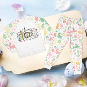 May include: White Easter-themed pajama set. The long-sleeved top features the text "true STORY he is risen" with a cross and floral design. The sleeves and pants are patterned with Easter eggs, bunnies, and flowers.