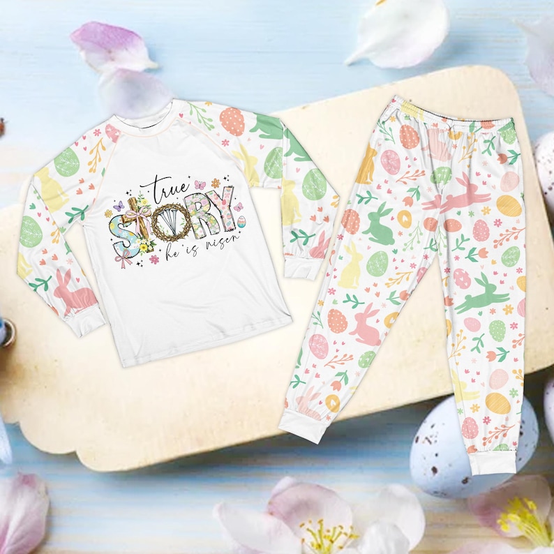 May include: White Easter-themed pajama set. The long-sleeved top features the text "true STORY he is risen" with a cross and floral design. The sleeves and pants are patterned with Easter eggs, bunnies, and flowers.