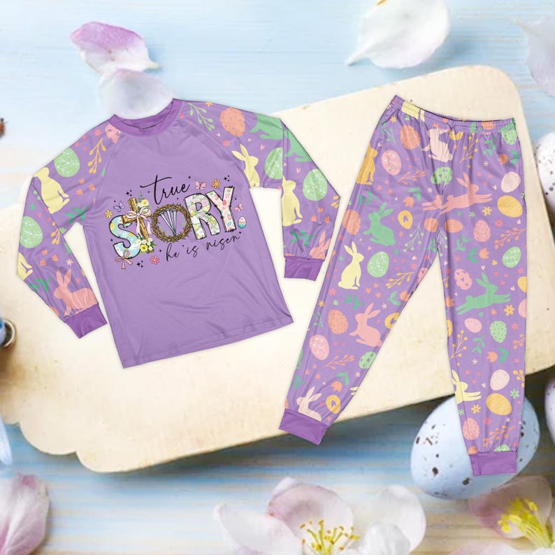 May include: A lavender pajama set featuring a long-sleeved top and matching pants. The top has the text "true STORY he is risen" with Easter-themed graphics. The pants and sleeves are decorated with Easter eggs and bunnies.