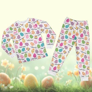May include: Easter-themed pajama set in white, featuring a pattern of multi-coloured eggs. Each egg has a unique design and the words "Custom name". The set includes a long-sleeved top and matching trousers.