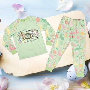May include: A light green Easter-themed pajama set. The long-sleeved shirt features the text "True Story, He is Risen" with floral and cross accents. The pajama pants have a pattern of Easter eggs, bunnies, and flowers. The set is made of a soft material.