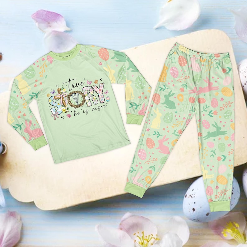 May include: A light green Easter-themed pajama set. The long-sleeved shirt features the text "True Story, He is Risen" with floral and cross accents. The pajama pants have a pattern of Easter eggs, bunnies, and flowers. The set is made of a soft material.