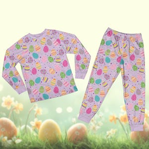 May include: A lilac pajama set with a long-sleeved top and trousers, patterned with colourful Easter eggs. The eggs are in various designs and colours, including blue, green, yellow, and pink. The pajamas have a relaxed fit and are made of a soft material.