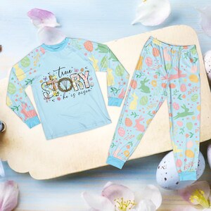 May include: Light blue Easter-themed pajama set. The long-sleeved top features the text "True Story, He is Risen" with a floral wreath design. The sleeves and pants are decorated with Easter eggs, bunnies, and floral patterns. The pajama set is made of a soft material.