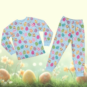 May include: A light blue pajama set with a repeating pattern of colourful Easter eggs. The long-sleeved top and trousers have a relaxed fit. The eggs are decorated with stripes, dots, and other designs in various colours.