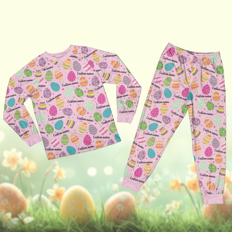 May include: A light pink pajama set with a long-sleeved top and trousers. The set is patterned with colourful Easter eggs and the text "Custom name". The pajamas are made of a soft material. The design is suitable for Easter.