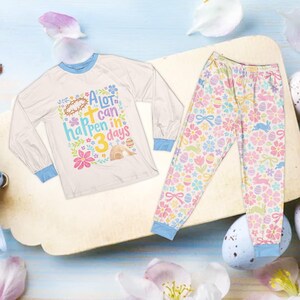 Op de afbeelding: Witte pyjama-top met lange mouwen en bijpassende broek. De top heeft blauwe randen en een kleurrijke afbeelding met de tekst "A lot can happen in 3 days". De broek heeft een bloemen- en Paas-thema patroon met konijntjes, eieren en strikken.