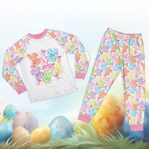 May include: Easter-themed pajama set featuring a white long-sleeved shirt with pastel bunny graphics and text, paired with matching pajama pants. The shirt has pink trim and the pants have a repeating bunny pattern. The text includes words like "Risen" and "Strong".