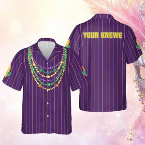 Peut inclure: Chemise violette à rayures verticales jaunes et vertes. L'avant présente un motif de collier de perles vertes, violettes et dorées. Le dos de la chemise porte l'inscription "YOUR KREWE" en jaune. La chemise a des manches courtes.