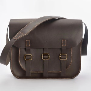 Puede incluir: Un bolso de mensajero de cuero marrón con dos correas ajustables y tres cierres de hebilla. El bolso tiene un cierre de solapa y un asa superior.