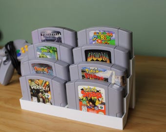 Può includere: Un supporto in plastica bianca contiene otto cartucce di gioco per Nintendo 64. Le cartucce sono disposte in due file da quattro. I titoli dei giochi visibili sono "Forsaken 64", "Turok", "Turok 2", "Quake II", "Super Mario 64", "Doom", "Pokemon Stadium" e "Pokemon Snap".