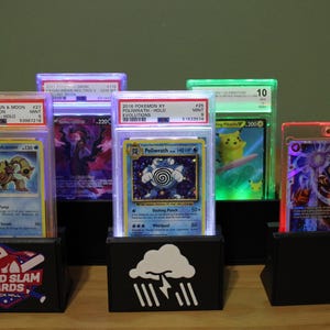 May include: A display of graded Pokémon trading cards in illuminated black stands. The cards feature various characters and are encased in protective holders. The stands have logos, including "Grand Slam Cards, Rock Hill."