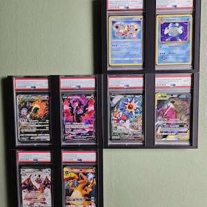May include: A wall display of nine Pokemon trading cards in protective cases. The cards are from different sets and feature various Pokemon characters, including Charizard, Volcanion, Poliwrath, and Starmie. The cards are in mint condition and are displayed in a grid pattern.