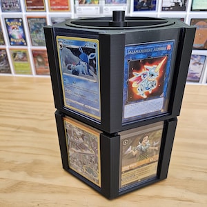 May include: A black hexagonal rotating display stand with six slots for displaying trading cards. The stand is filled with various trading cards, including a Salamangreat Almiraj card, a Glacion card, a Show White card, and a card with a picture of a building.